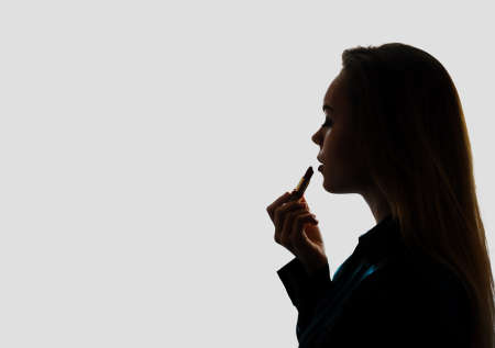 Close up portrait of attractive girl rouging her lips. She is holding lipstick. Her mouth is gently open. Isolated on grey backgroundの写真素材