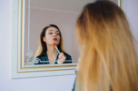 portrait in mirror of sexy smiling young woman model with glamour red lips, bright makeup, perfect clean skin. white teeth, applying dry cosmetic tonal foundation on the face using makeup brush.の写真素材