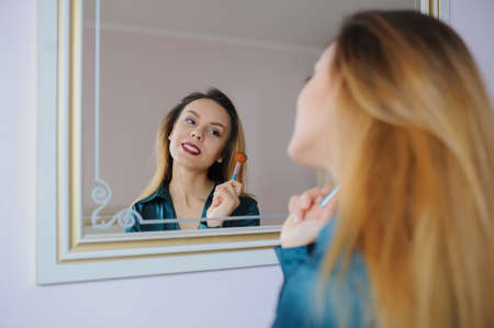 portrait in mirror of sexy smiling young woman model with glamour red lips, bright makeup, perfect clean skin. white teeth, applying dry cosmetic tonal foundation on the face using makeup brush.の写真素材