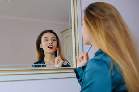 portrait in mirror of sexy smiling young woman model with glamour red lips, bright makeup, perfect clean skin. white teeth, applying dry cosmetic tonal foundation on the face using makeup brush.の写真素材
