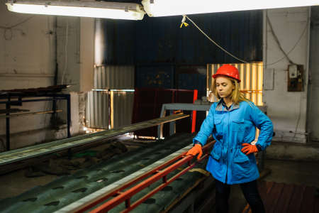 Portret of nice beautiful woman in red safety helmet at metal tile roof manufacturing factory examine the forming processの写真素材