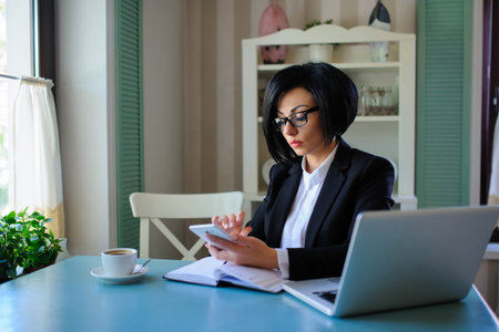 business lady with glasses dressed in black suit working on a laptop ( portable compute ) and have a cup of coffee on the table; beautiful woman is browsing internet; handsome girl with notebook;の写真素材