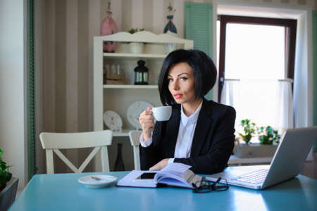 business lady dressed in black suit working on a laptop ( portable compute ) and have a cup of coffee on the table; beautiful woman is browsing internet; handsome girl with notebook;の写真素材