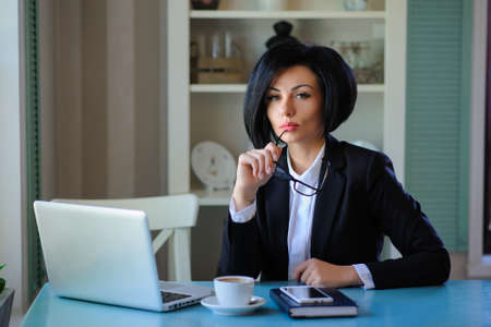 business lady with glasses dressed in black suit working on a laptop ( portable compute ) and have a cup of coffee on the table; beautiful woman is browsing internet; handsome girl with notebook;の写真素材
