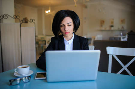 business lady dressed in black suit working on a laptop ( portable compute ) and have a cup of coffee on the table; beautiful woman is browsing internet; handsome girl with notebook;の写真素材