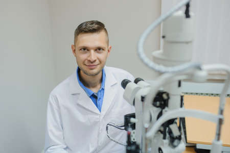 Young beautiful doctor/eye specialist/optometrist in an ophthalmologic clinic sitting on the chair near the biomicroscop with slit lamp and expect the patient.の写真素材