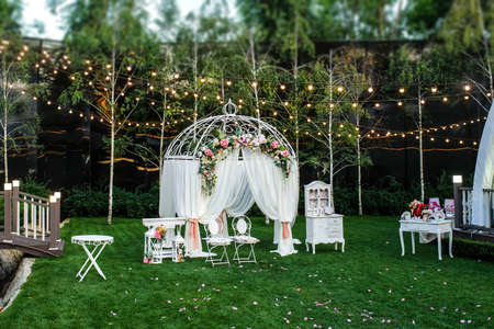 White arch for the wedding ceremony decorated with roses, ranunculus and peony. Vintage furniture elements: chair, table, lights bulbsの写真素材