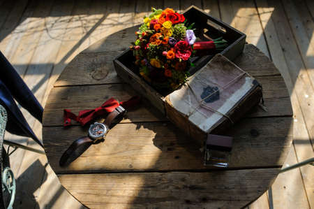 The groom's watch and red bowtie and his perfume with the bride's bouquet of flowers on the wooden tableの写真素材