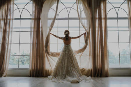 Beautiful bride in long white dress looks out the window.の写真素材