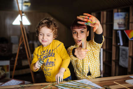 Young mother and her son playing with color paints in art studio. Dirty hands with color on them hold the brushes and laughの写真素材