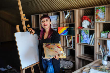 Close up portrait of beautiful female artist with purple hair and dirty hands with different paints on them, holding paintbrushes and laugh in her art studio.の写真素材