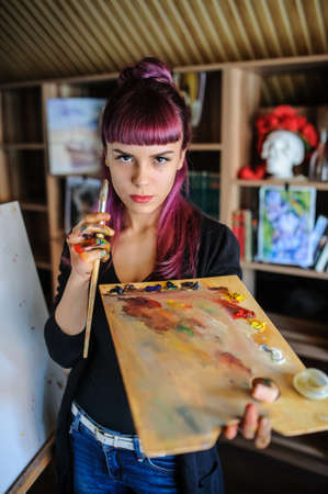Close up portrait of beautiful female artist with purple hair and dirty hands with different paints on them, holding paint brushes near her face and eyes.の写真素材