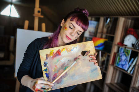 Close up portrait of beautiful female artist with purple hair and dirty hands with different paints on them, holding paint brushes near her face and eyes.の写真素材