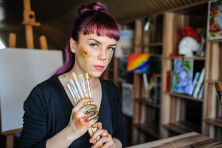 Beautiful and serious female artist with purple hair and dirty hands with different paints on them, holding paintbrushes in her art studio.の写真素材
