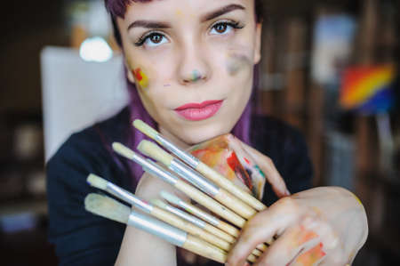 Close up portrait of beautiful female artist with purple hair and dirty hands with different paints on them, holding paint brushes near her face and eyes.の写真素材