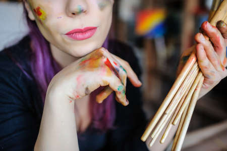 Crop picture of lips and face of beautiful female artist with purple hair and dirty hands with different paints on them, holding paintbrushes in her art studio.の写真素材