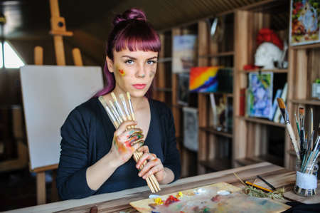 Beautiful and serious female artist with purple hair and dirty hands with different paints on them, holding paintbrushes in her art studio.の写真素材