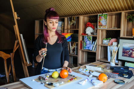 Beautiful and serious female artist with purple hair and dirty hands with different paints on them, holding paintbrushes in her art studio.の写真素材
