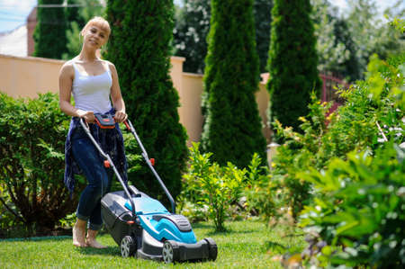 Young handsome  gardener girl is cutting the grass with the mowerの写真素材