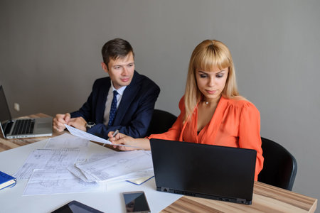 A business woman and man at a wooden office desk  looking at the project on the paper and computer...