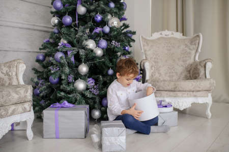 a 6-year blond boy with a present in his hand near a christmas tree with purple toys and two white vintage armchairsの写真素材