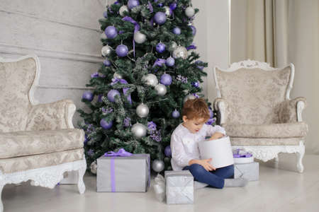 a 6-year blond boy with a present in his hand near a christmas tree with purple toys and two white vintage armchairsの写真素材