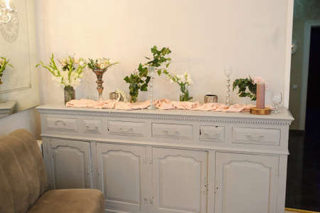 A white large vintage sidebord in a wedding restaurant hall with vases with flowers, candles and peach curtains on it next to a beige sofaの写真素材