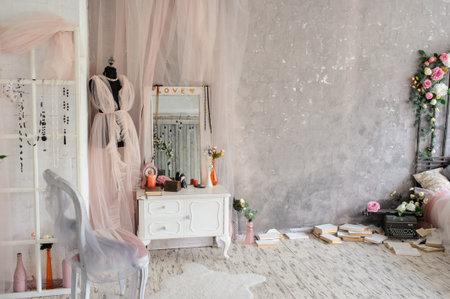 Lateral view of a vintage room in a photo studio with a white vintage wooden dressing table with a big mirror, vases with flowers, a black mannequin with a dress from pink curtain on it next to a chair and a bed surrounded by booksの写真素材
