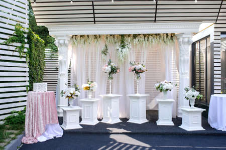 A beautiful white rectangular wedding arch for welcoming guests decoreted with flowers and curtains surrounded by white pillars with vases with flowers on them along the arch and two tablesの写真素材