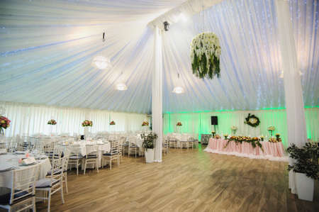 The interior of a wedding banquet hall: round tables with white tablecloth and all necessary supplies for dinner on them, bouquet of flowers; white curtains, beige floor, white chairs, presidium table with pink tableclothの写真素材