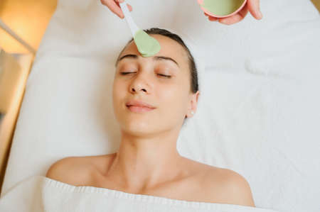 Close up head shot of a Woman having a green mask applied. Therapist applying the mask on the skin and forehead.の写真素材