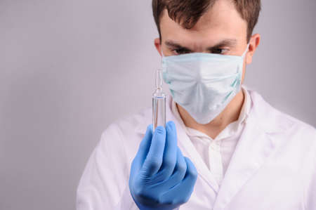 a doctor with mask in a white uniform looking at the ampoule with medicine that he is holding in his hand in a blue gloveの写真素材