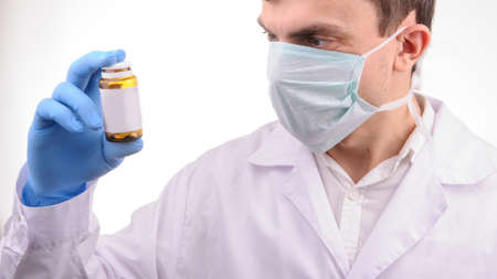 a doctor with mask in a white uniform looking at a bottle with pills that he is holding in his hand in a blue gloveの写真素材