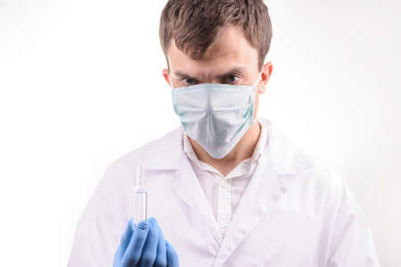 a doctor with mask in a white uniform looking at the ampoule with medicine that he is holding in his hand in a blue gloveの写真素材