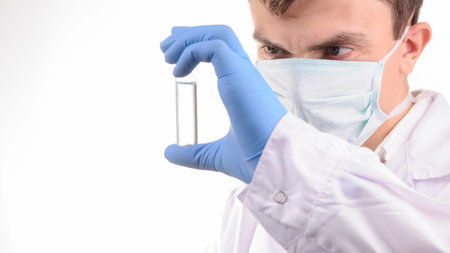 a doctor with mask in a white uniform looking at the ampoule with medicine that he is holding in his...