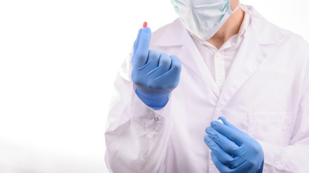 a doctor with mask in a white uniform looking at a red capsule that he is holding in his hand in a...