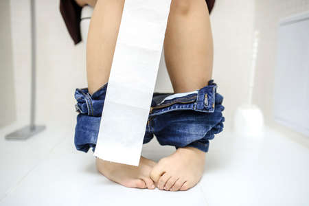 close up photo woman with constipation or diarrhoea sitting on toilet with her pants down arround her legs and a lot of toilet paper rolls on the floorの写真素材