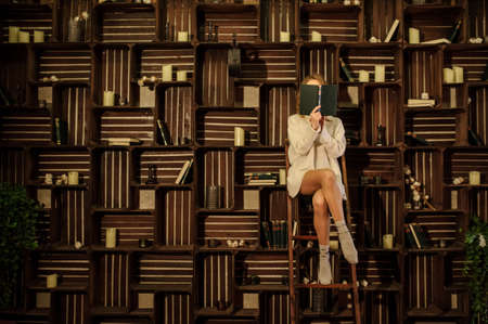 close up photo of a young woman in warm wool sweater and socks standing on the stairs reading a book next to the bookshelfの写真素材