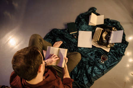 top view of man with a book in his hands sitting near a vintage typewriter on a dark blue velvet fabric on the floor with sheets of paper a glass of wine and books arroundの写真素材