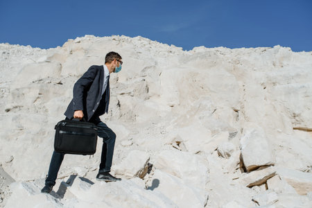 Young man businessman with a briefcase and face mask is climbing in the mountain. Concept of...