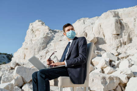 Young man businessman with a face mask is sitting on a chair and working on a notebook computer in the mountain in Coronavirus Covid 19 Pandemic. Concept of online work in coronavirus COVID 19 pandemic. Work from everywhereの写真素材