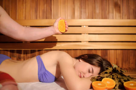 lateral view of a beautiful woman enjoying massage procedure with oranges and birch broom in a wooden saunaの写真素材