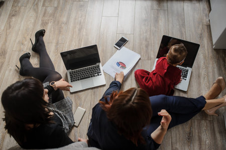 top view of two women in office outfit working on computers and discussing a report on the floor...