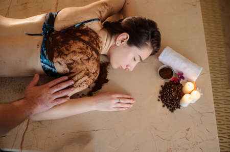 lateral view of a beautiful woman in spa salon having spa therapy with coffee done by a masseurの写真素材