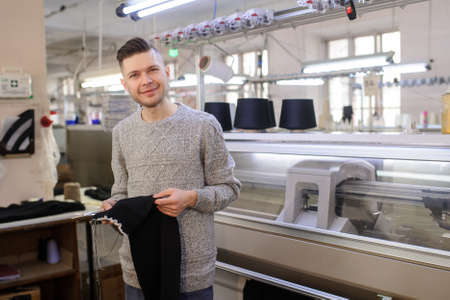 a young man analyzing a knitted piece of cloths near industrial knitted machines with black thread in conesの写真素材