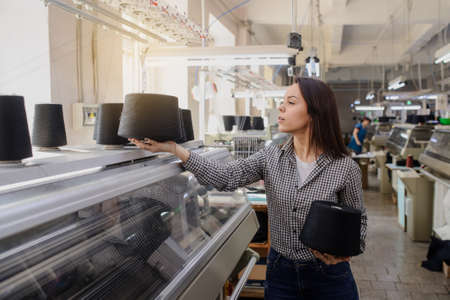 close up photo of a young woman with black threads in cones in her hands near industrial knitting machinesの写真素材