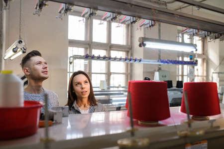 close up photo of a young woman and man looking and discussing near a industrial knitting machine with red cones on itの写真素材