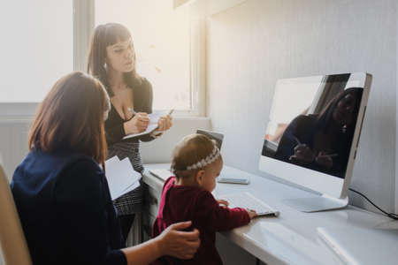 close up photo of two women in office outfit working on computers and discussing a report; one of them is with a baby girl in her armsの写真素材