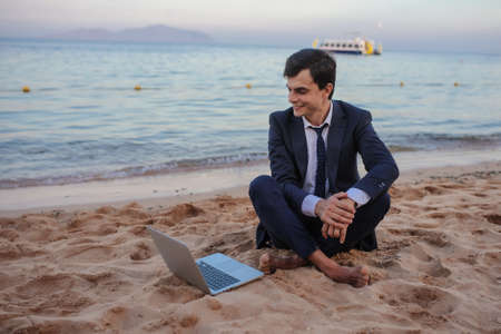 lateral view of a young man in suit with laptop sitting on the beach working on the seasideの写真素材