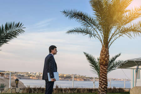lateral view of a young man in suit with laptop working on green grass near a resort on the seasideの写真素材